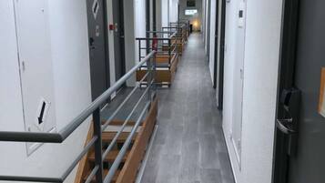 Un corredor largo y estrecho con una escalera de madera a la izquierda, barandillas de metal y un suelo de baldosas grises.