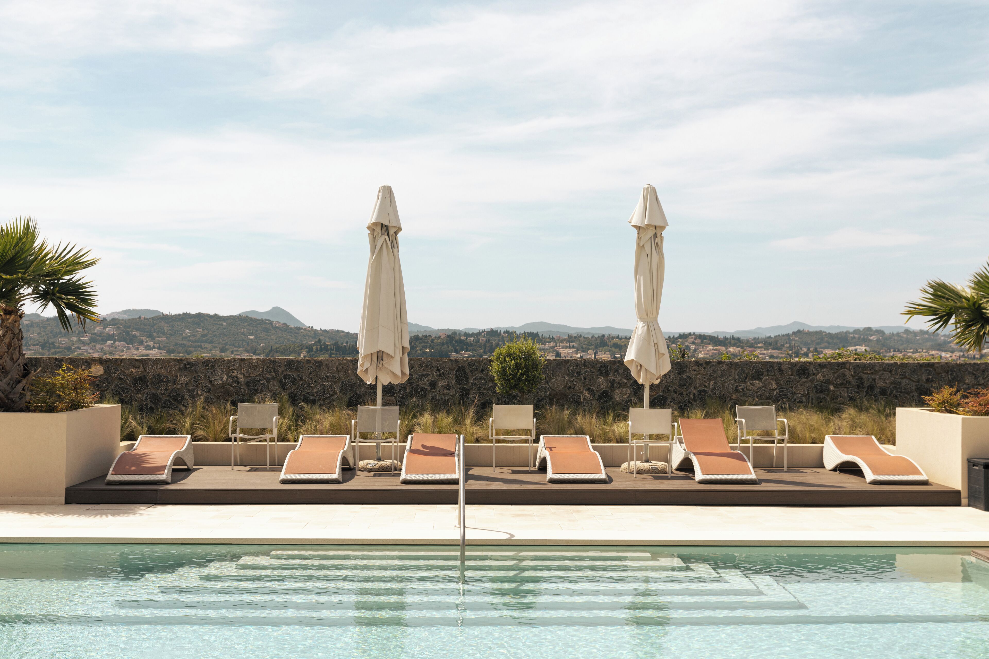 Ein Poolbereich mit Liegestühlen und Sonnenschirmen, eine Steinmauer und Blick auf ferne Hügel.
