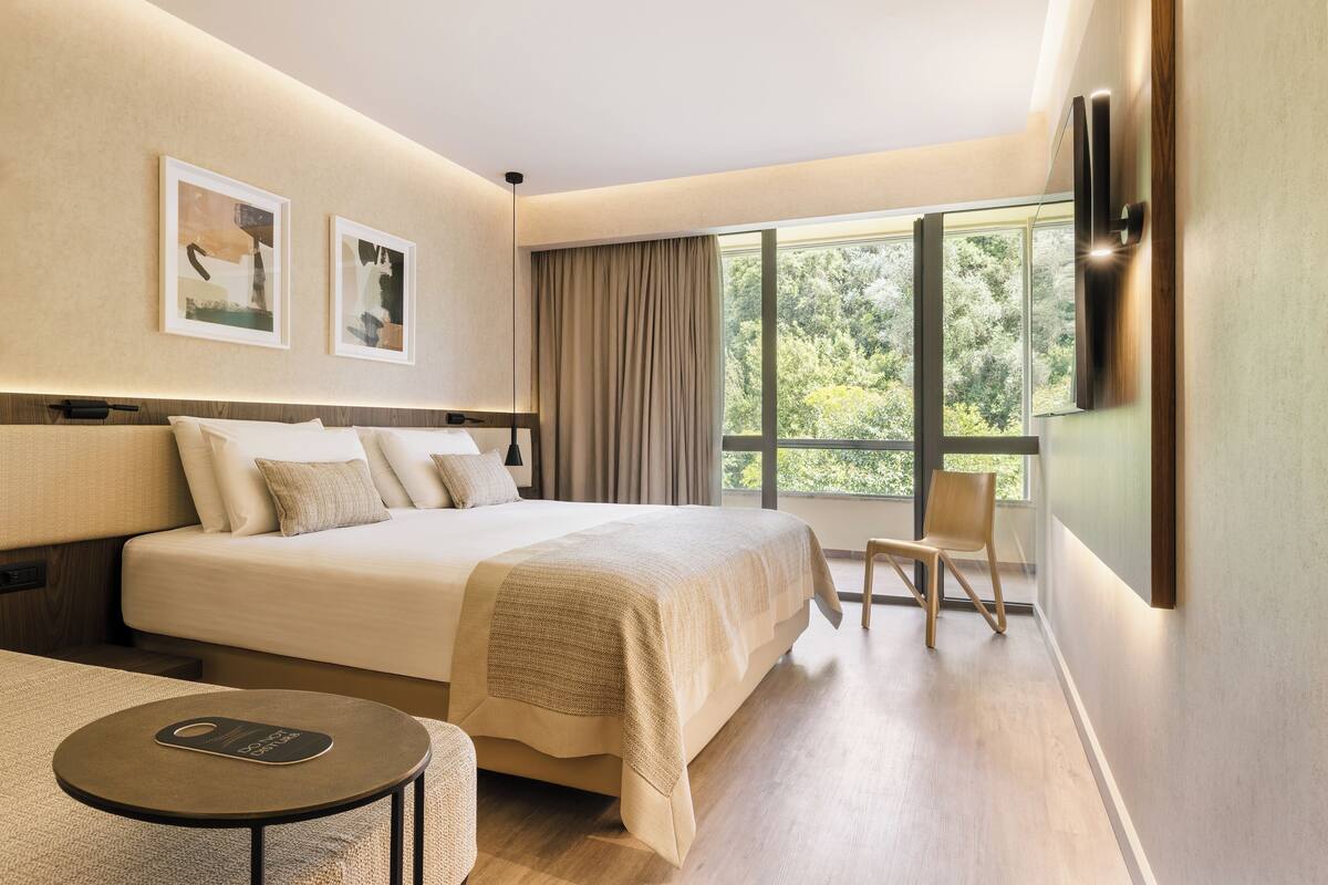 A modern hotel room with a large bed, a small table, a chair, and a large window offering a view of greenery.