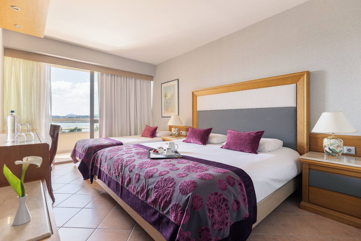 A hotel room with two beds, a desk, a chair, and a view of the sea.