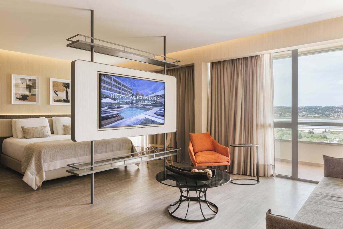 A modern hotel room with a bed, a TV mounted on a wall, a round table, and a large window with a view.
