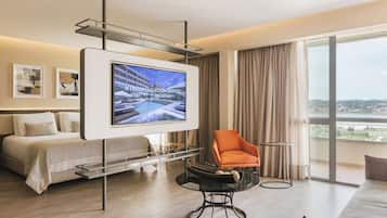 A modern hotel room with a bed, a TV mounted on a wall, a round table, and a large window with a view.