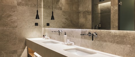 A modern bathroom with two sinks, a wooden bench, and pendant lights.