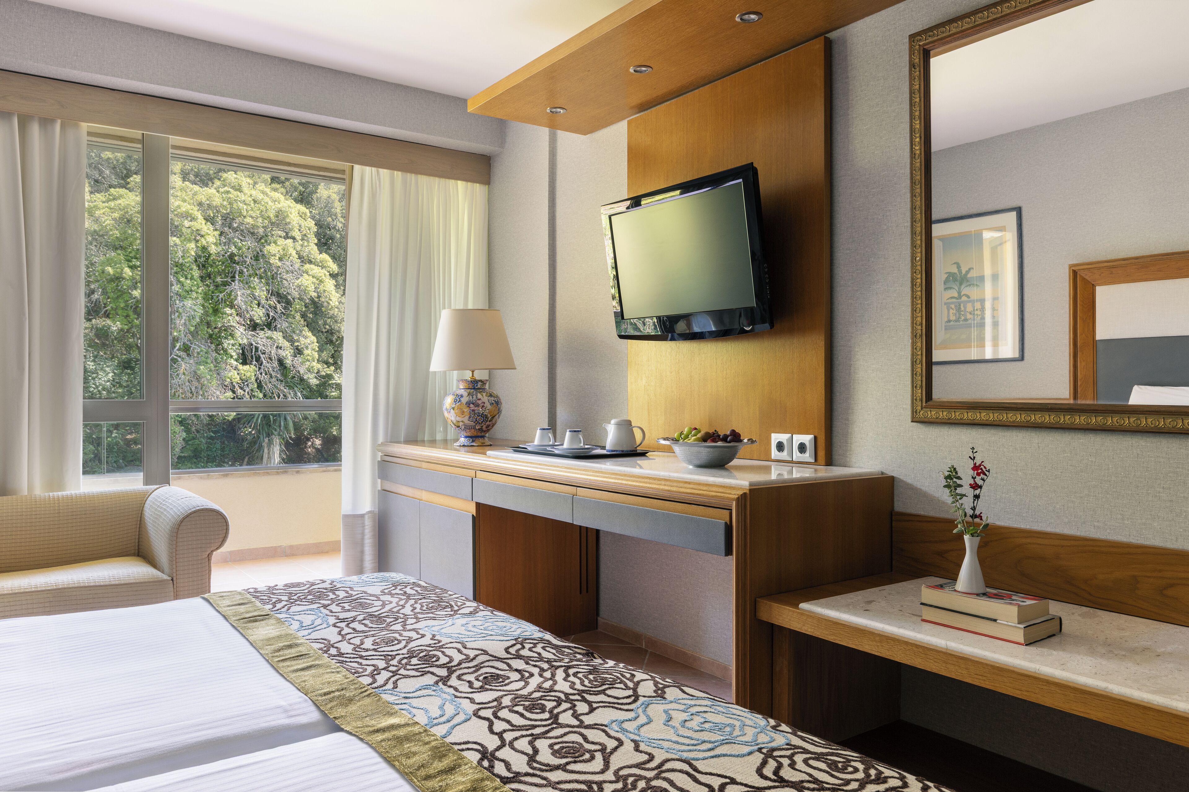 Habitación de hotel con cama, escritorio con televisor, ventana con cortinas y una mesita con un jarrón y libros.