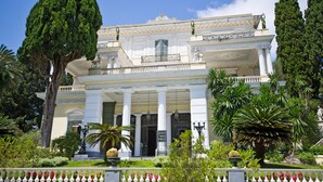 A grand building with classical architecture, featuring columns, statues, and a well-maintained garden with various plants and a decorative fence. - Divani Corfu Palace (Corfu)