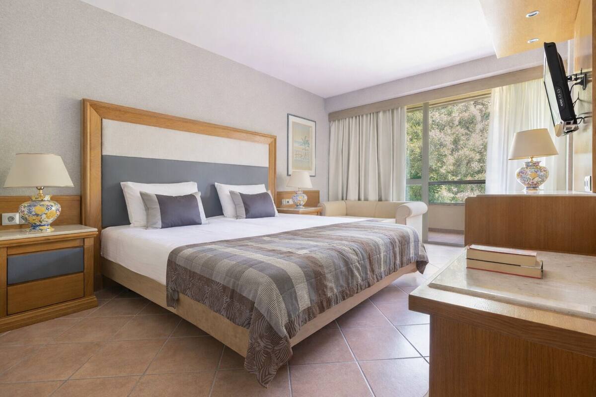 A hotel room with a bed, bedside tables, a desk, and a window.