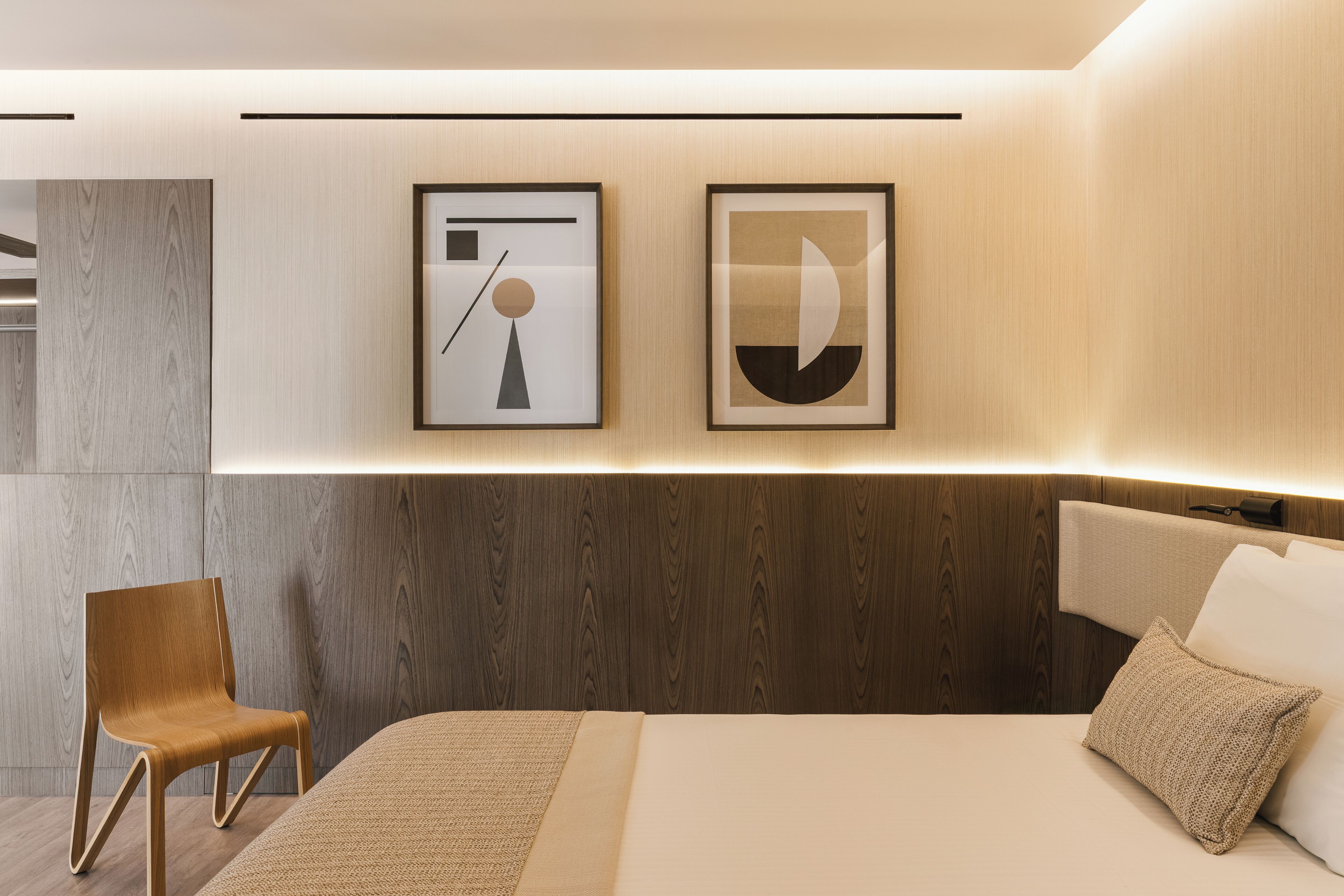 A modern hotel room with a wooden headboard, a bed with white linens, a brown chair, and two framed abstract artworks on the wall.