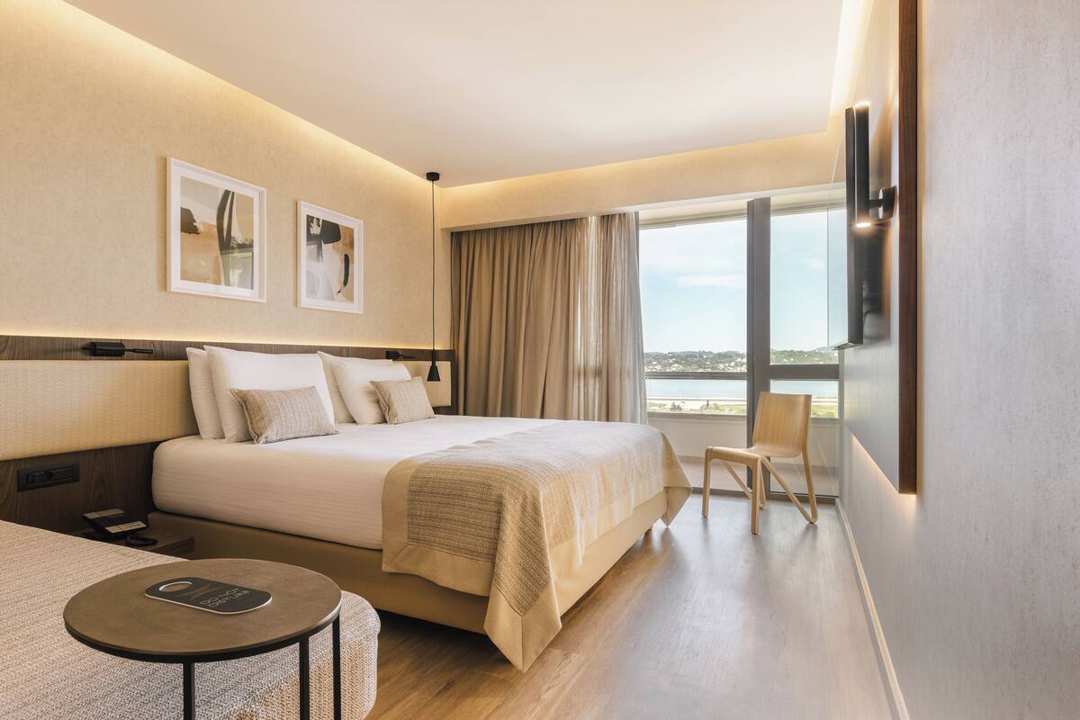 A hotel room with a bed, a small table, a chair, a TV, and a window with a view.