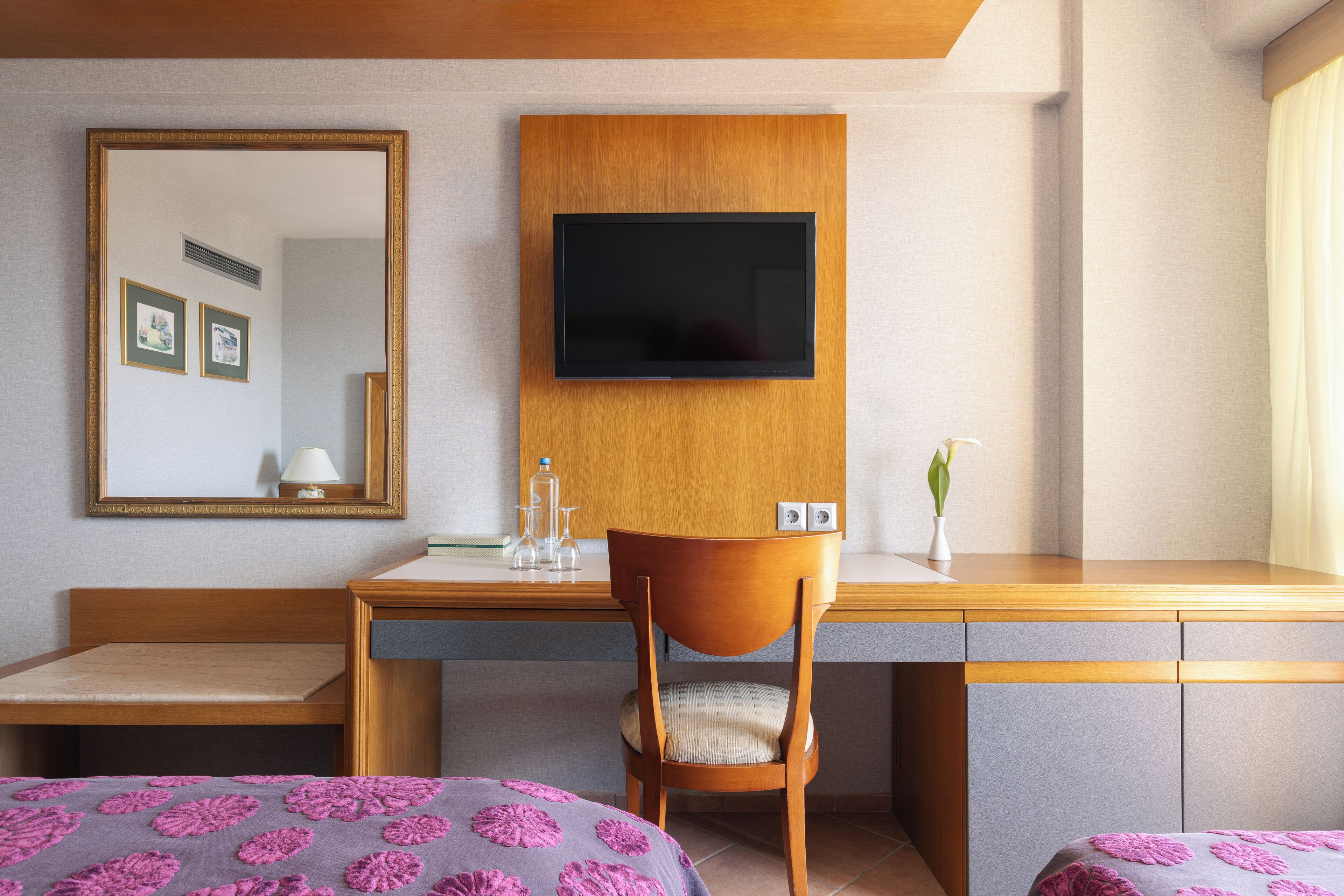 A hotel room with a wooden desk, a television mounted on the wall, a chair, a bed with a purple floral bedspread, and a mirror.