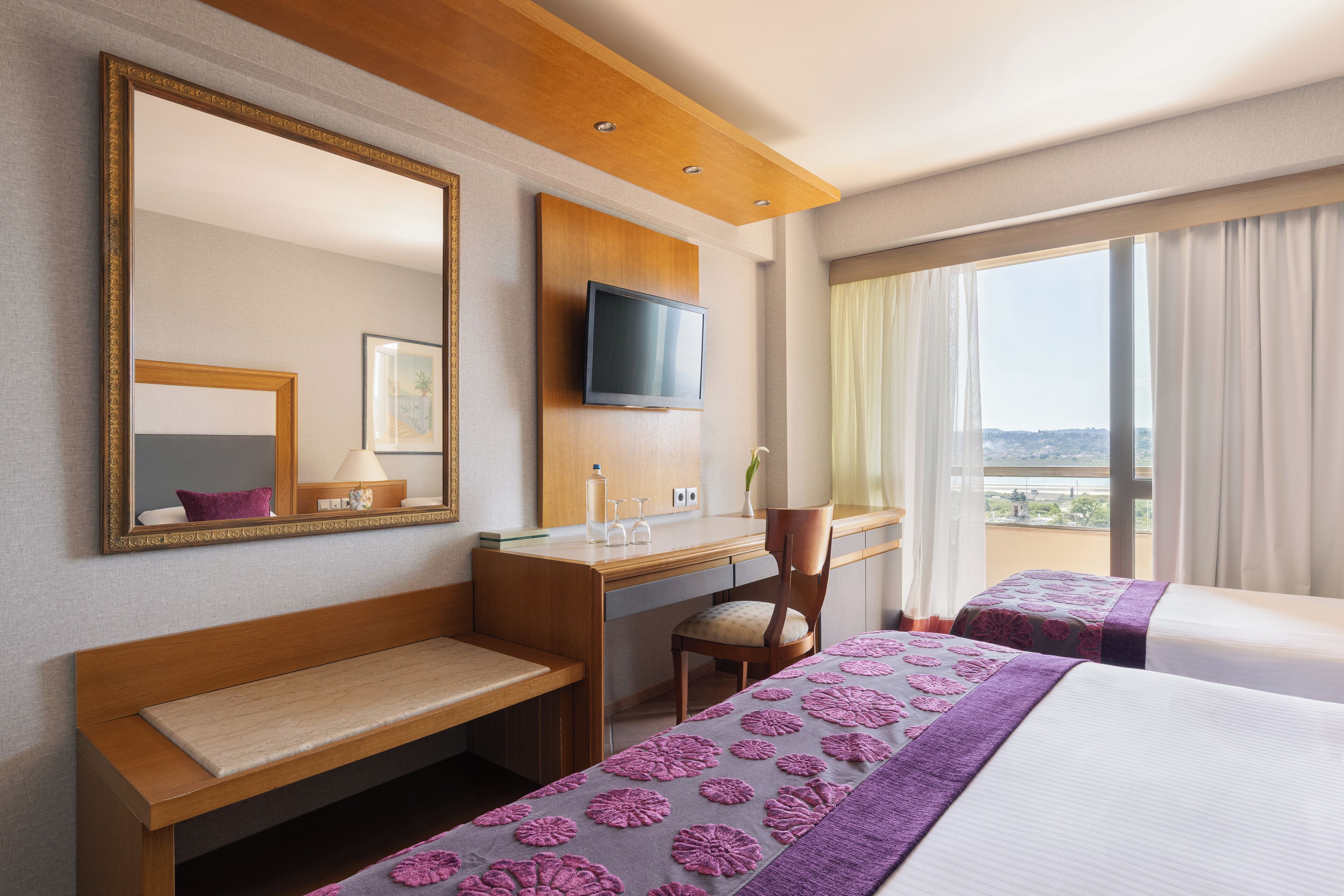 Ein Hotelzimmer mit Schreibtisch aus Holz, einem an der Wand montierten Fernseher, einem großen Spiegel, einem Bett mit violett gemustertem Bettbezug und Blick auf die Stadt durch das Fenster.