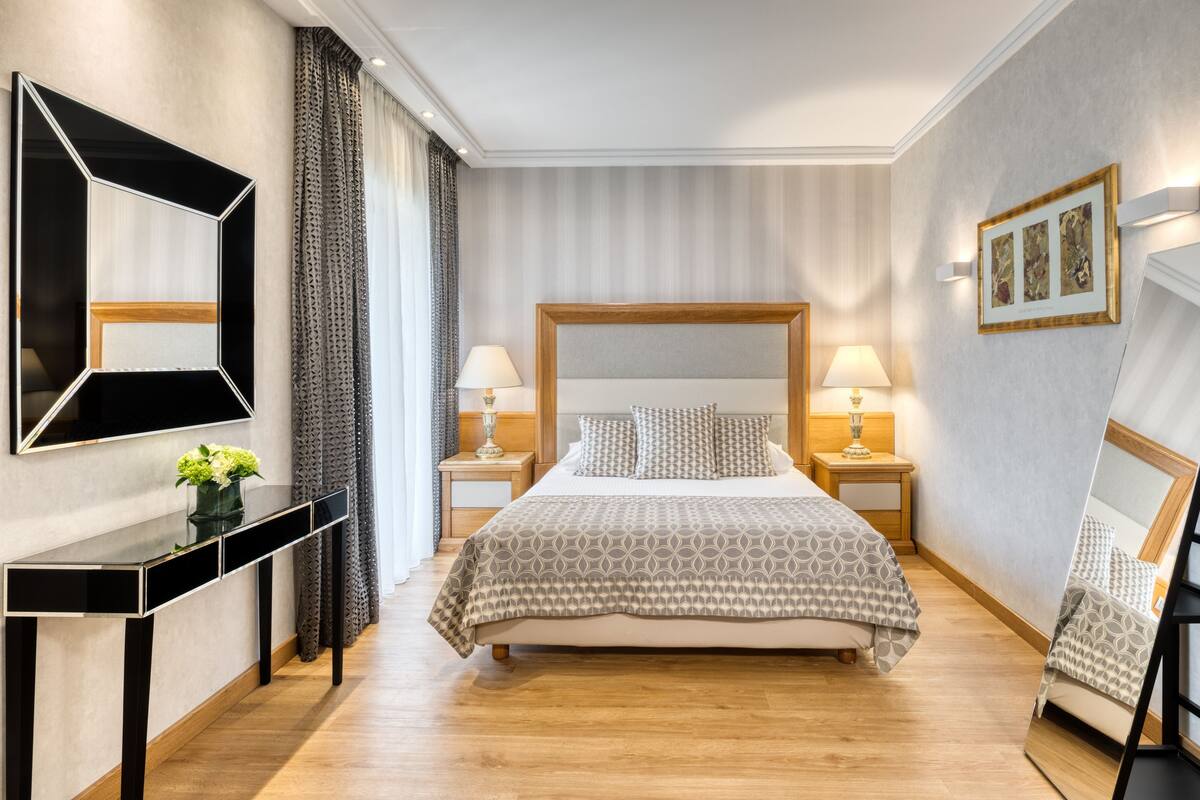 A modern hotel room with a large bed, bedside tables with lamps, a console table with a vase, and a large mirror.