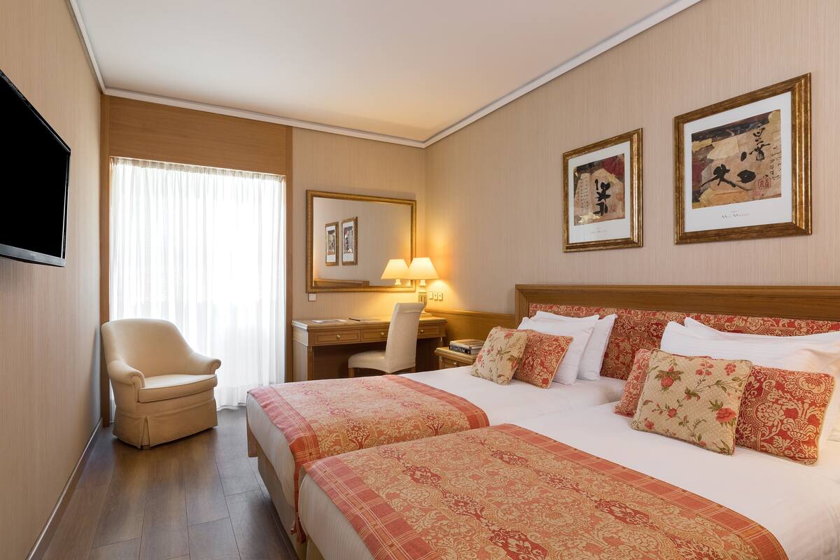 A hotel room with two beds, a desk, a chair, a TV, and artwork on the walls.