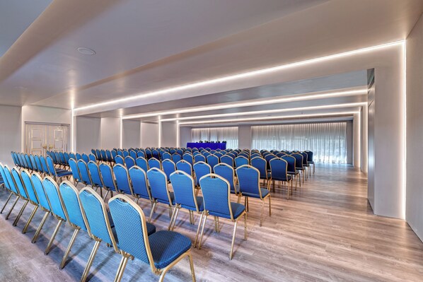 A conference room with rows of blue chairs, a wooden floor, and a long table at the front.