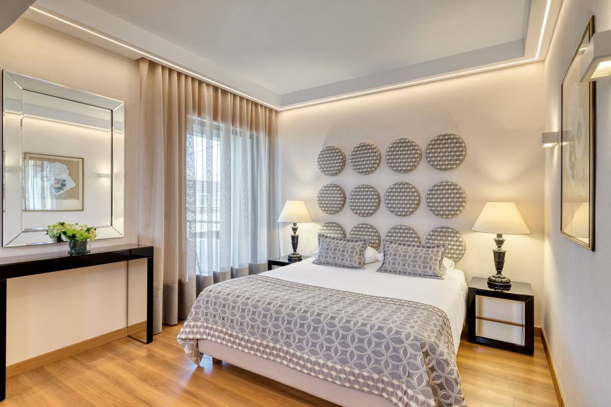 A modern hotel room with a large bed, a bedside table with a lamp, a mirror, and decorative wall art.