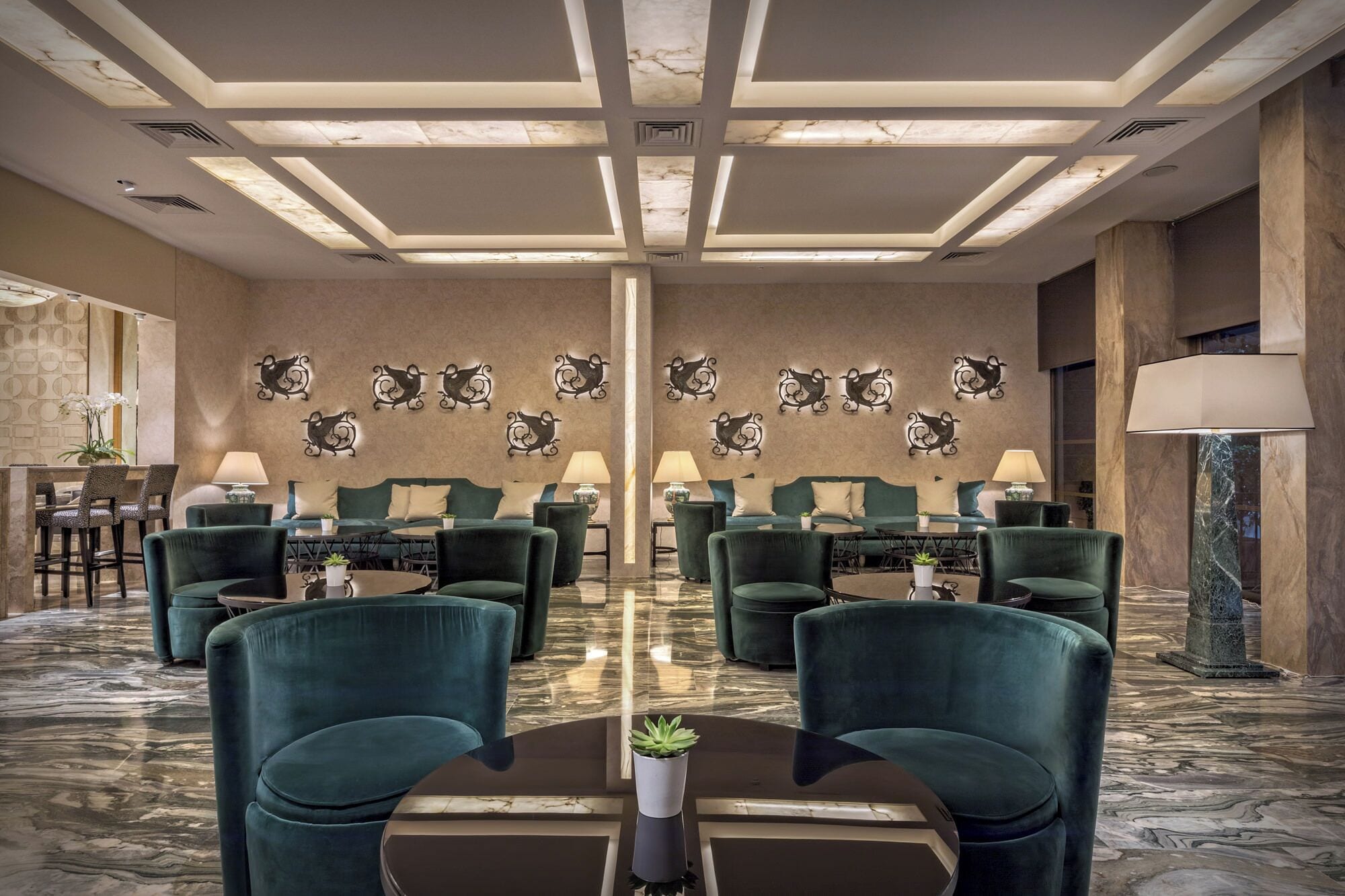 A hotel lobby with a marble floor, green upholstered seating, and wall-mounted decorative elements.