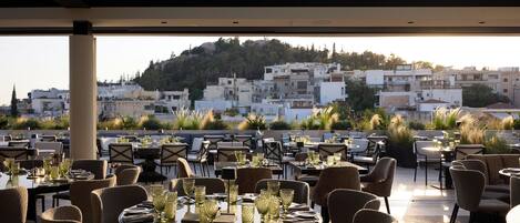 An outdoor dining area with tables set for a meal, overlooking a hillside with buildings.