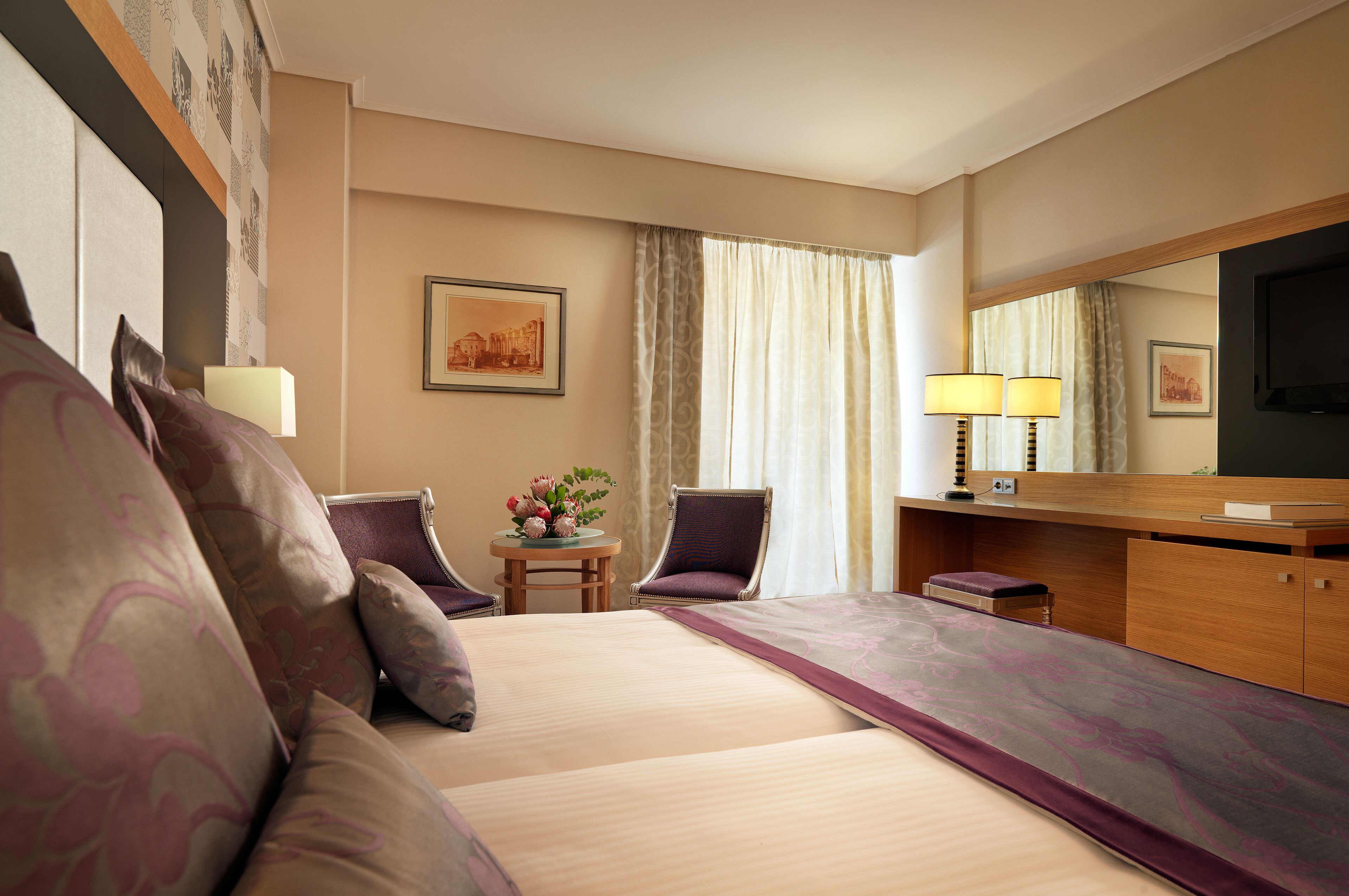 Une chambre d’hôtel avec un grand lit, deux fauteuils, une petite table ornée d’un vase de fleurs, un bureau avec une lampe et un téléviseur.