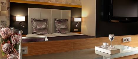 A hotel room with a bed, bedside lamps, a flat-screen TV, a glass coffee table, a vase with flowers, and a chair.