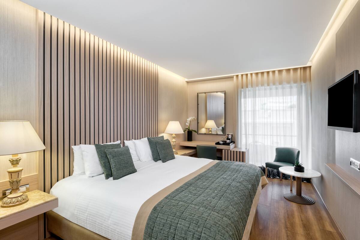 A hotel room with a large bed, bedside tables, a desk, a chair, a TV, and a window with curtains.