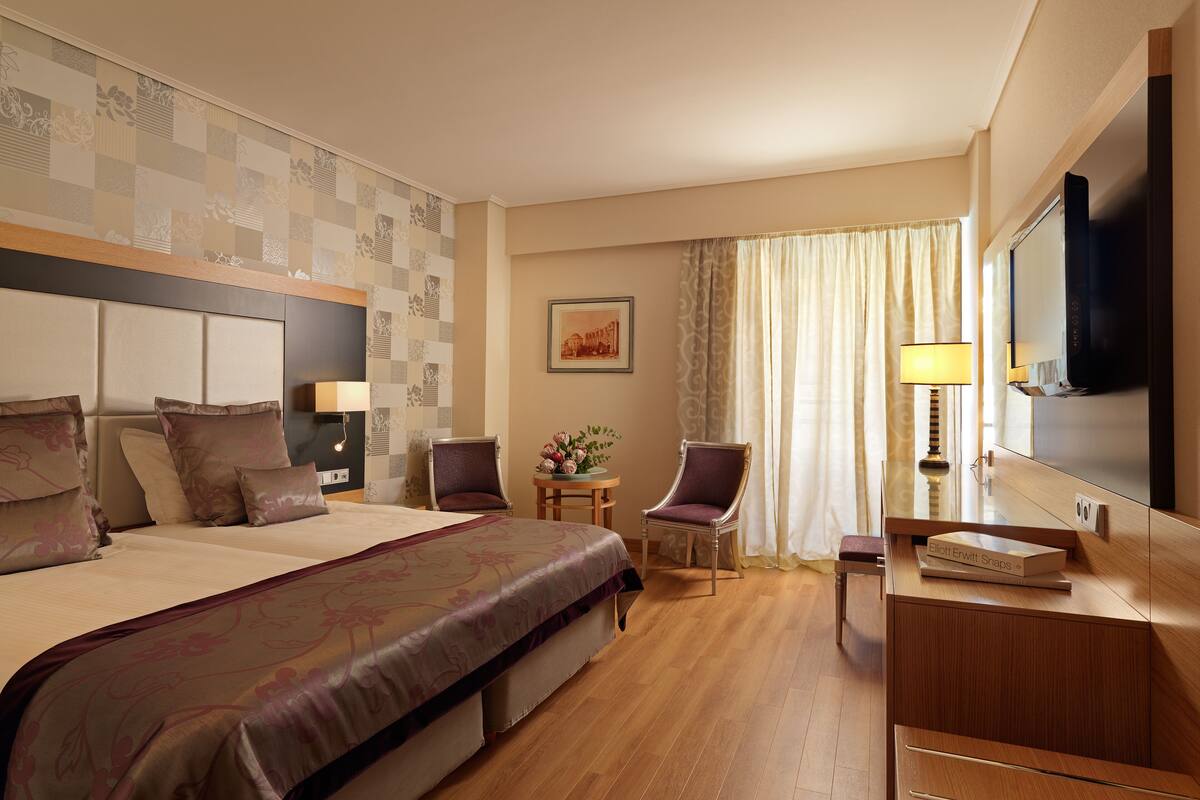 A hotel room with a large bed, two chairs, a small table with flowers, a TV, and a bookshelf.
