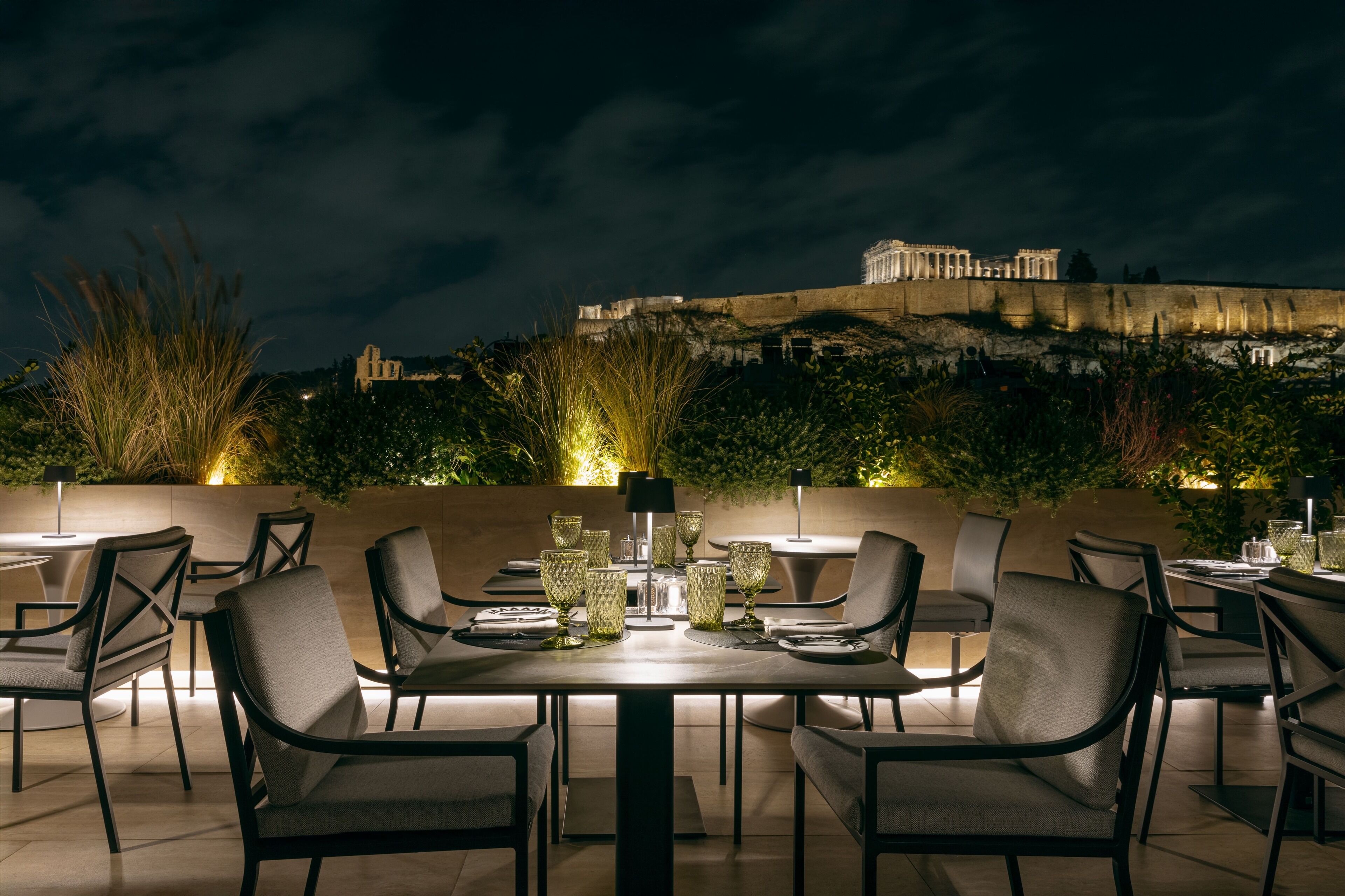 An outdoor dining area with tables and chairs, a view of ancient ruins in the background, and a well-lit ambiance.