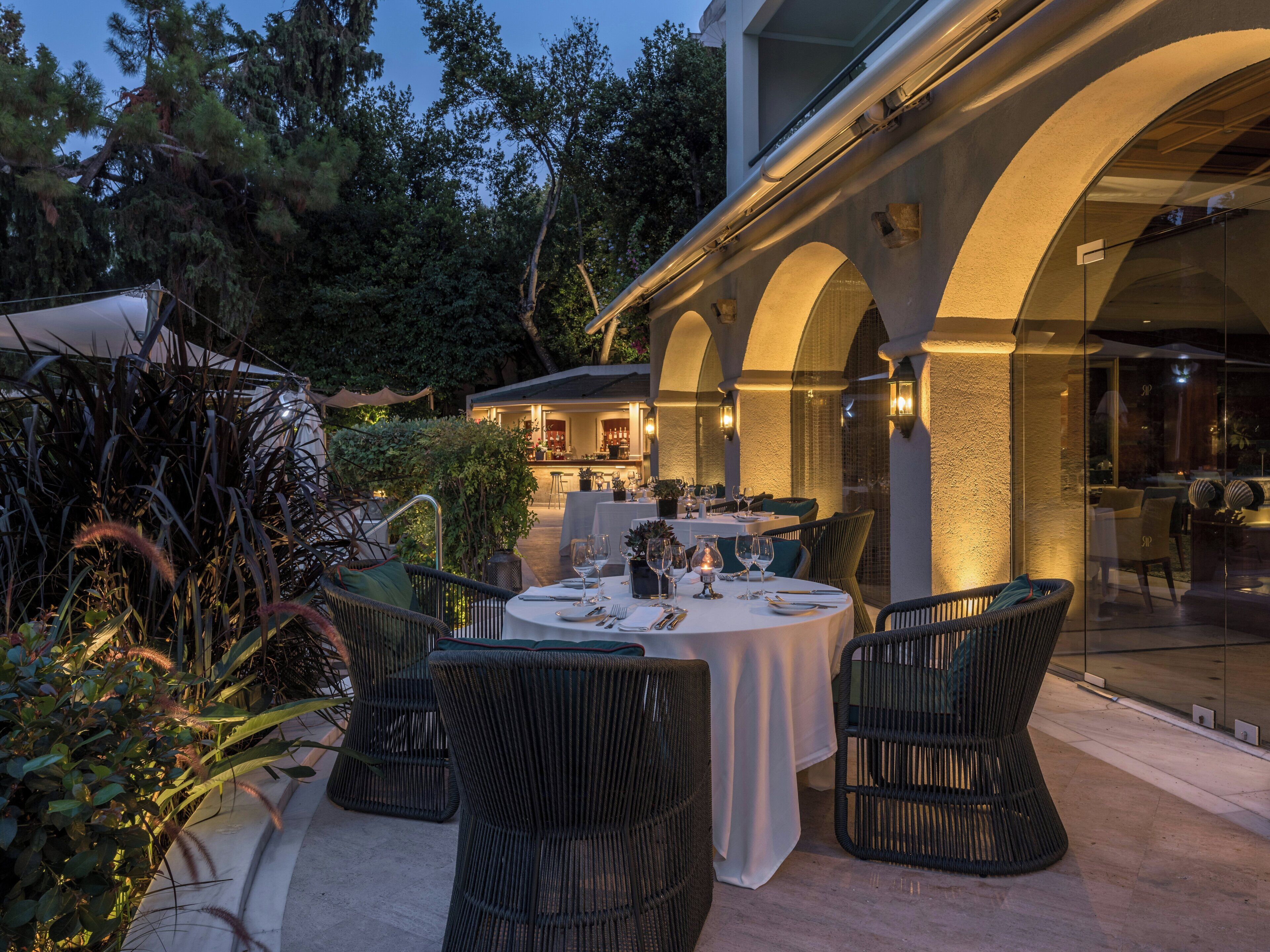 An outdoor dining area with round tables and wicker chairs, surrounded by greenery and a covered section with a bar.