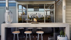 Un  área de bar al aire libre moderna con mostrador de hormigón, taburetes metálicos y una variedad de botellas y vasos.