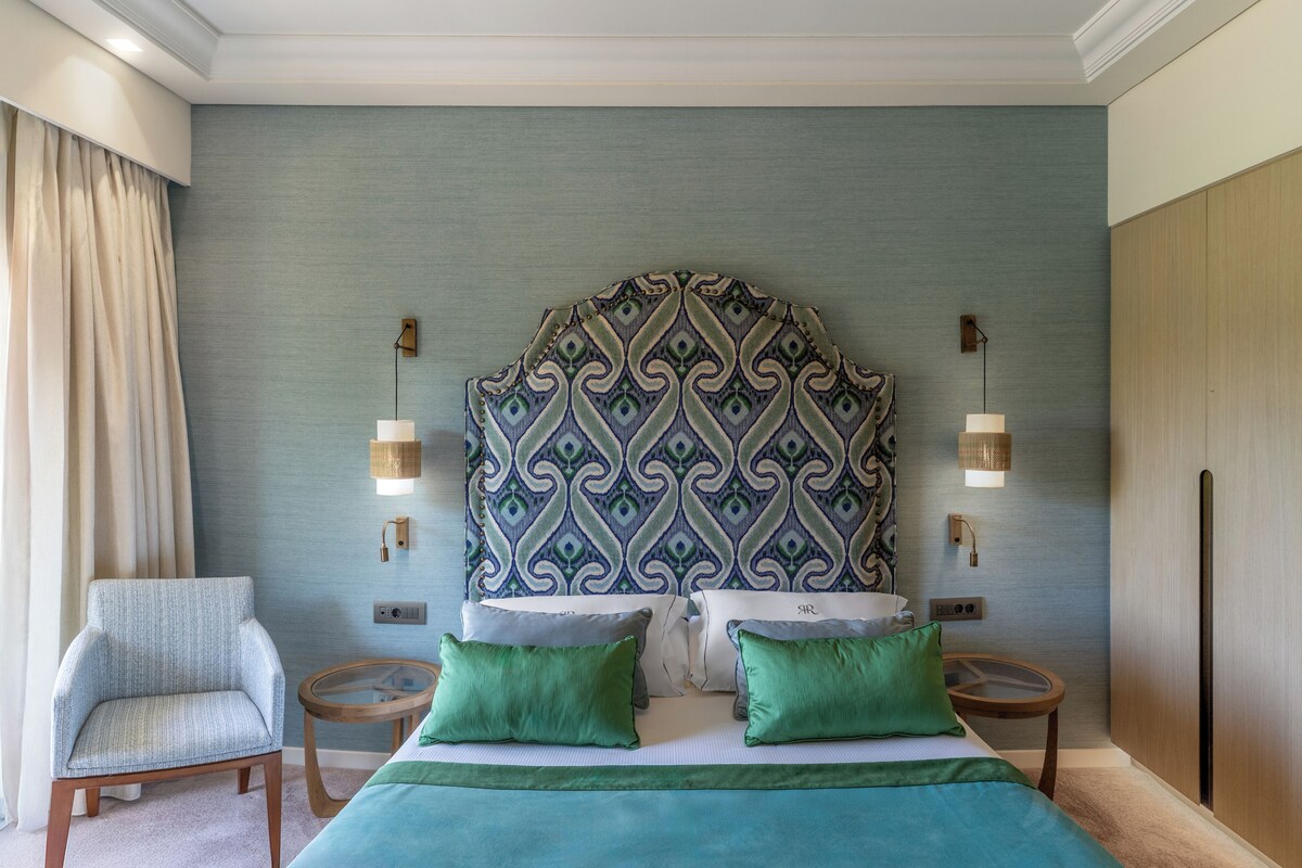 A hotel room with a large bed, two bedside tables, a chair, and a patterned headboard.