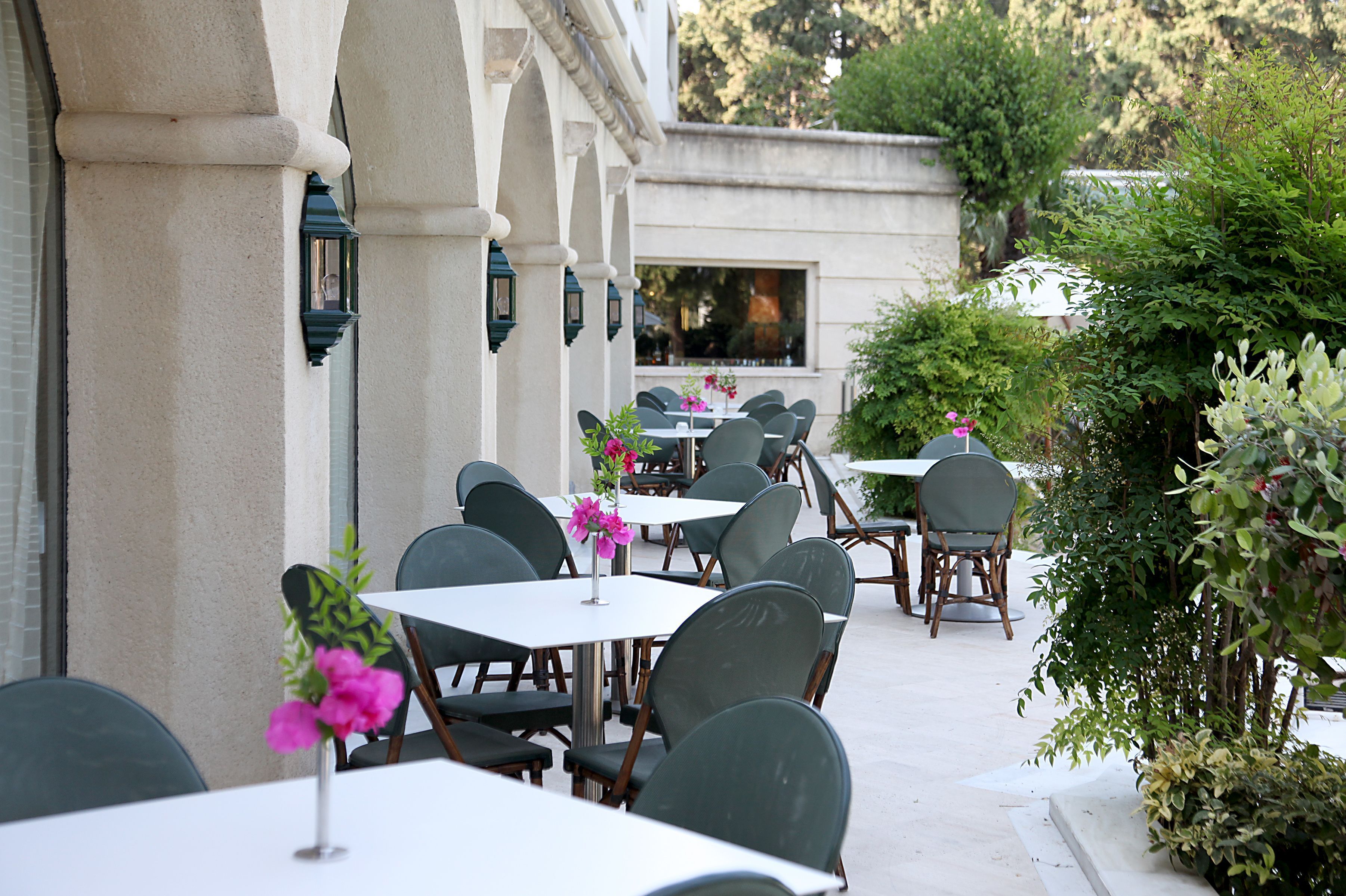 An outdoor dining area with tables and chairs, surrounded by greenery and a building facade.