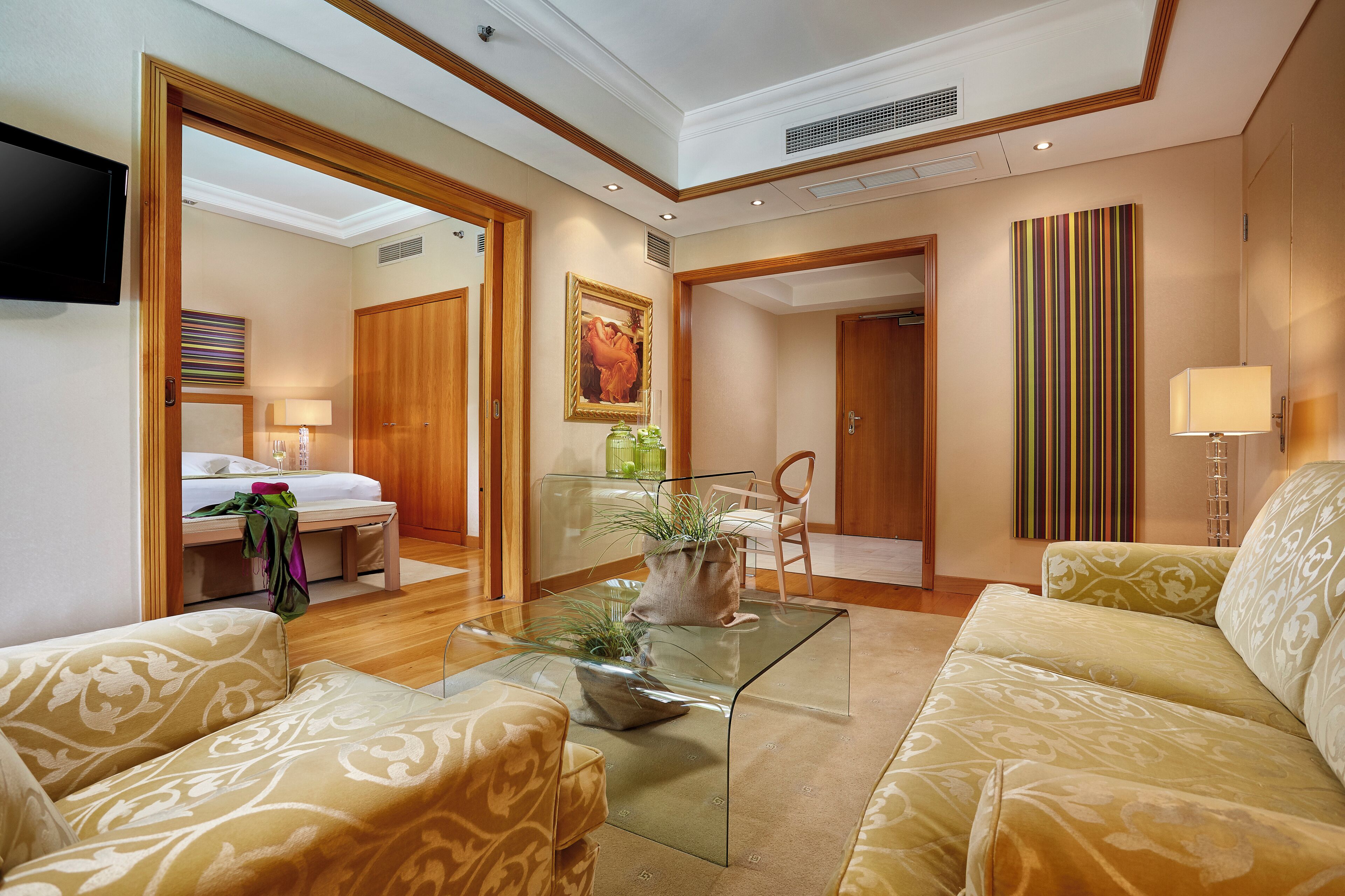 A hotel room with a glass coffee table, two patterned sofas, a bed, a TV, a painting, and a striped wall decoration.