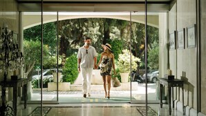 Una pareja caminando por la entrada de un hotel con puerta de vidrio, vistas al exterior con vegetación y suelo de baldosas.