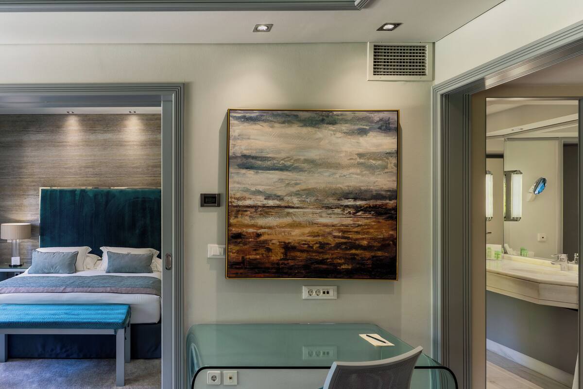 A modern hotel room with a bed, bedside table, lamp, and a large landscape painting.