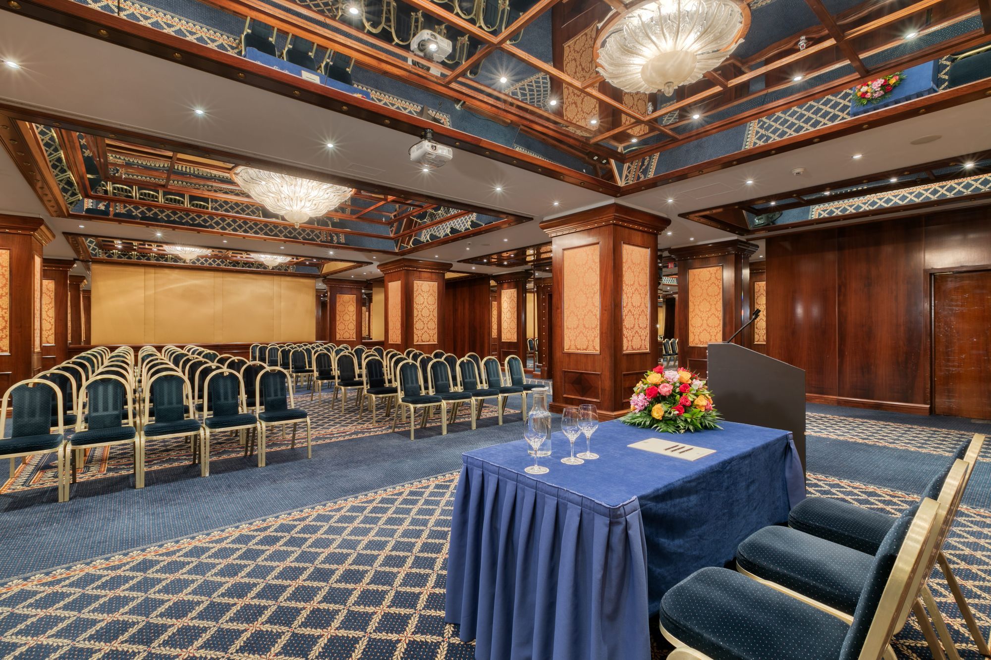 Uma sala de conferências com fileiras de cadeiras, uma mesa com toalha azul e um arranjo floral.