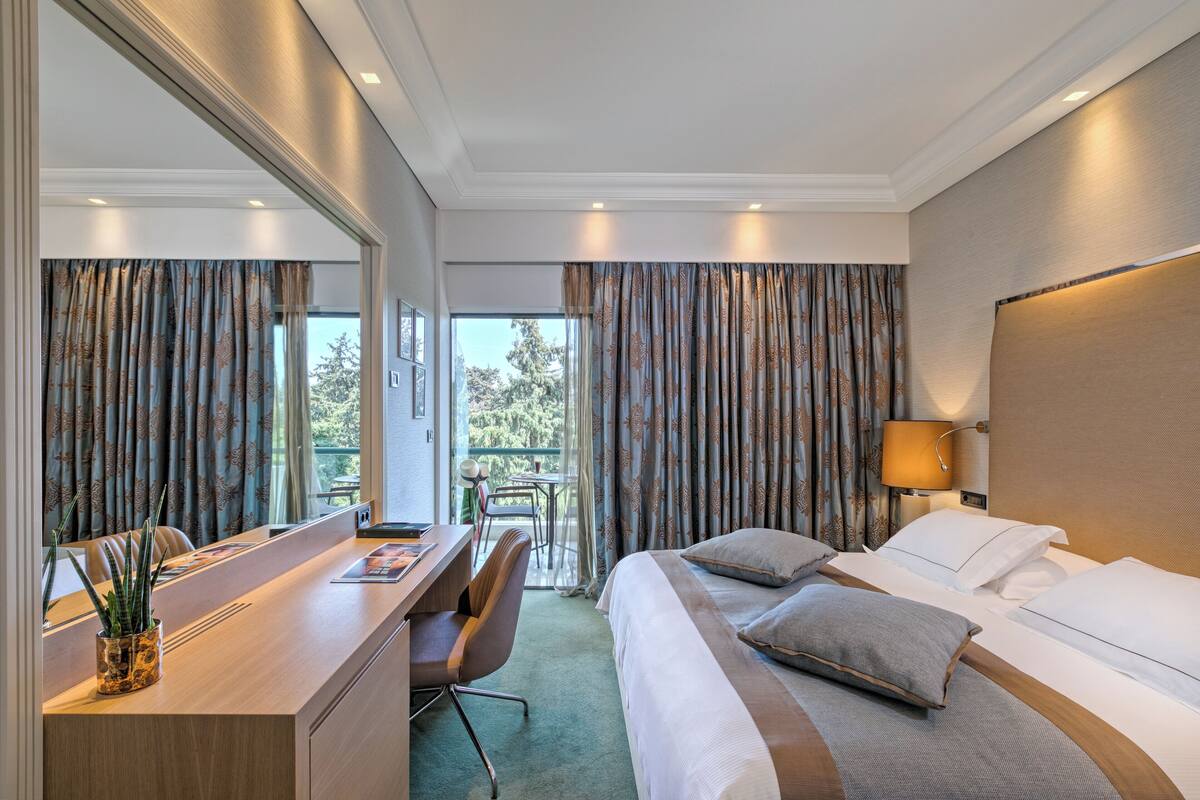A hotel room with a bed, bedside table, desk, chair, and a view of the outdoors.