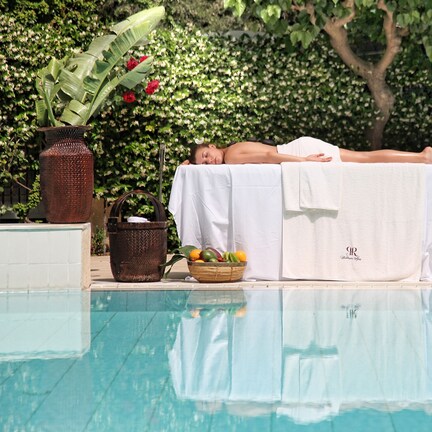 A person lying on a white towel by a pool, with a wicker basket of fruit and a vase of flowers nearby.