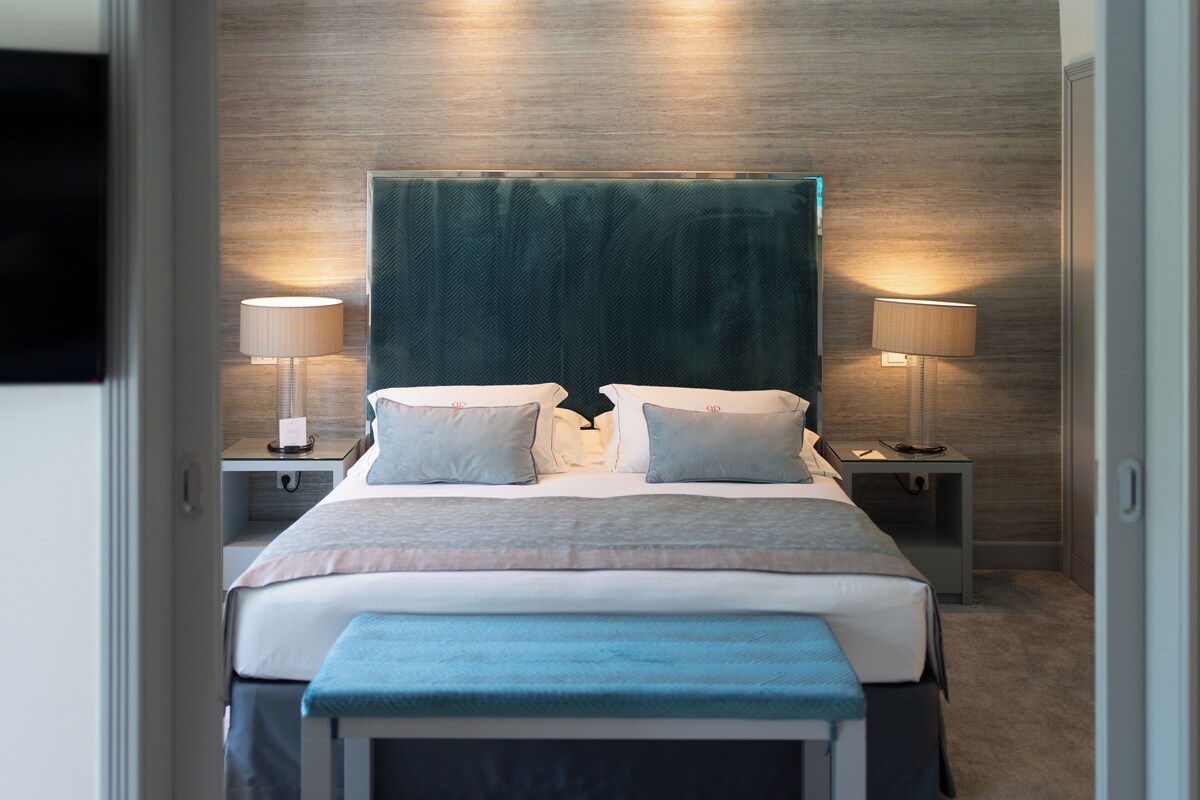 A modern hotel room with a large bed, two bedside tables with lamps, a bench at the foot of the bed, and a wooden wall.