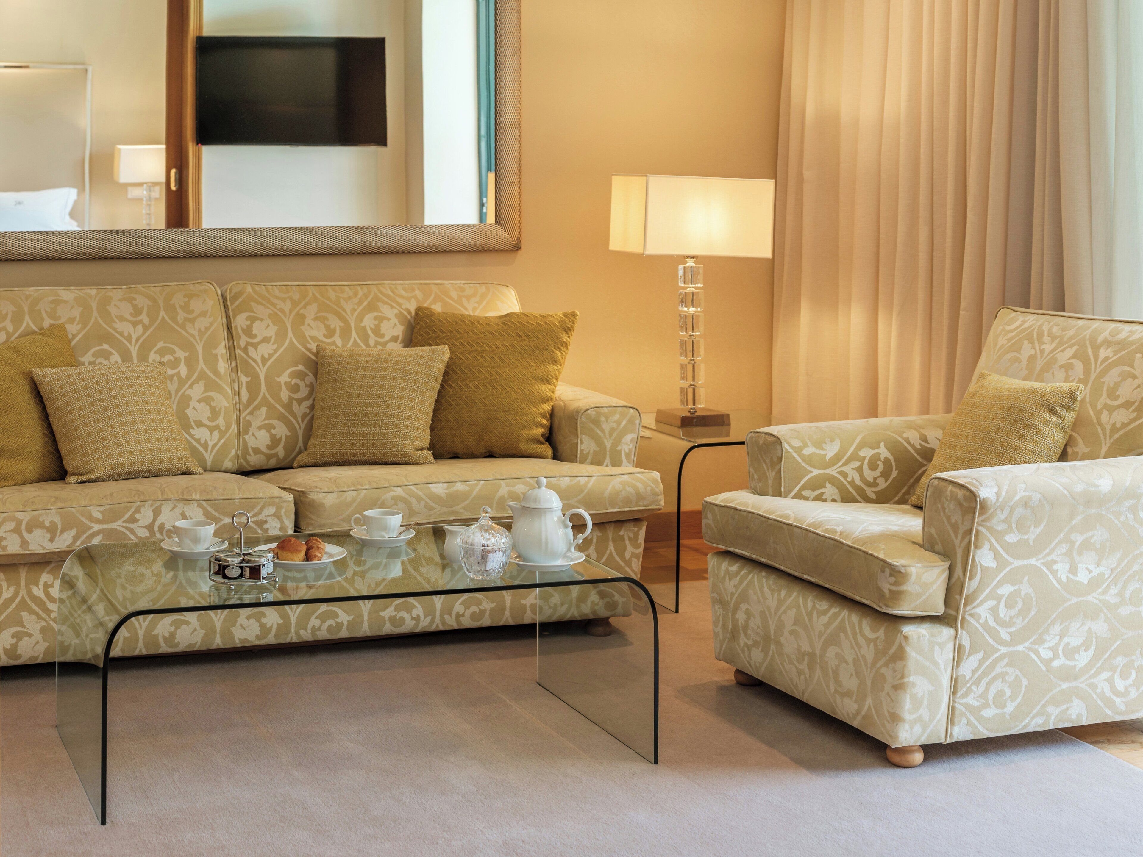 A living room with a sofa, armchair, and coffee table. There is a lamp and a television on the wall.