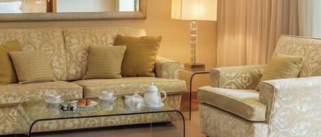 A living room with a sofa, armchair, and coffee table. There is a lamp and a television on the wall.