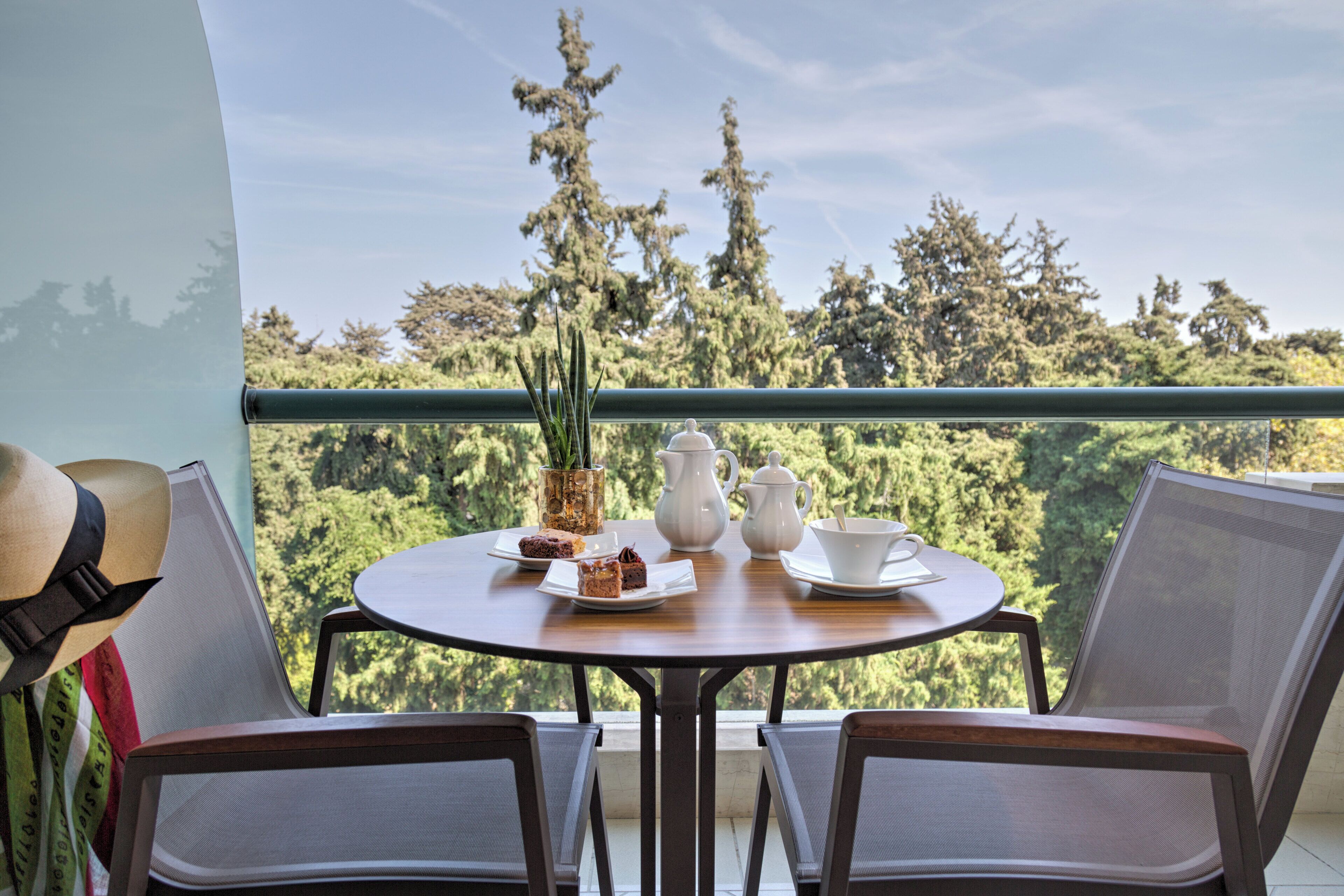 Et rundt bord med to stole, en vase med grønne planter, en hvid kande, en tepotte og en kop på en balkon med udsigt over en skov.