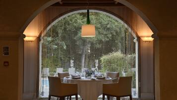 A dining area with a round table set for four, surrounded by chairs, under a large arched window with a mesh screen.