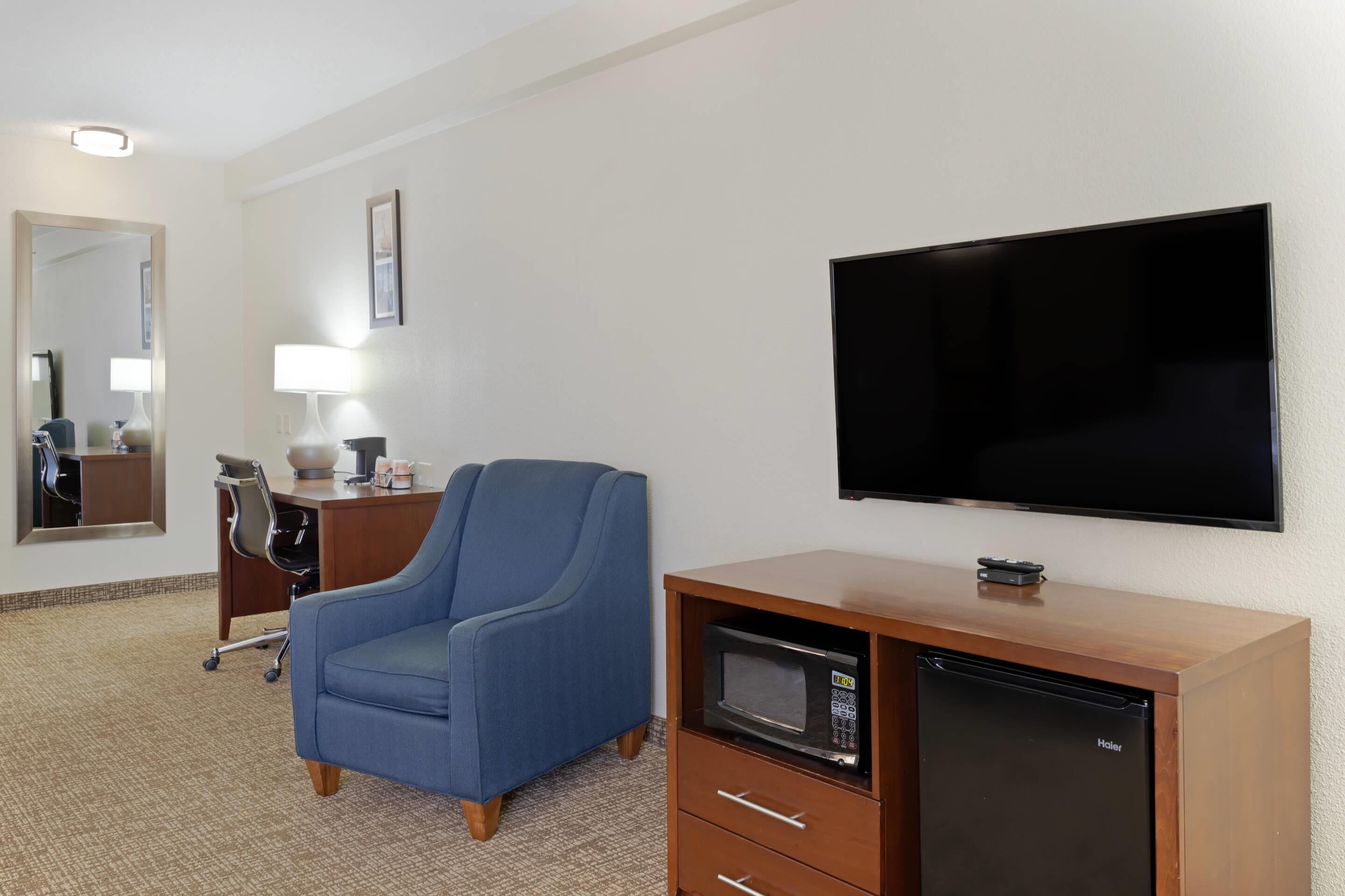 A hotel room with a flat-screen TV, a microwave, a blue armchair, a desk with a lamp, and a mirror.