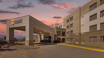 Entrada de hotel con zona techada, estacionamiento y fachada del edificio.