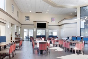A spacious dining area with multiple tables and chairs, a flat-screen TV mounted on the wall, and large windows allowing natural light.