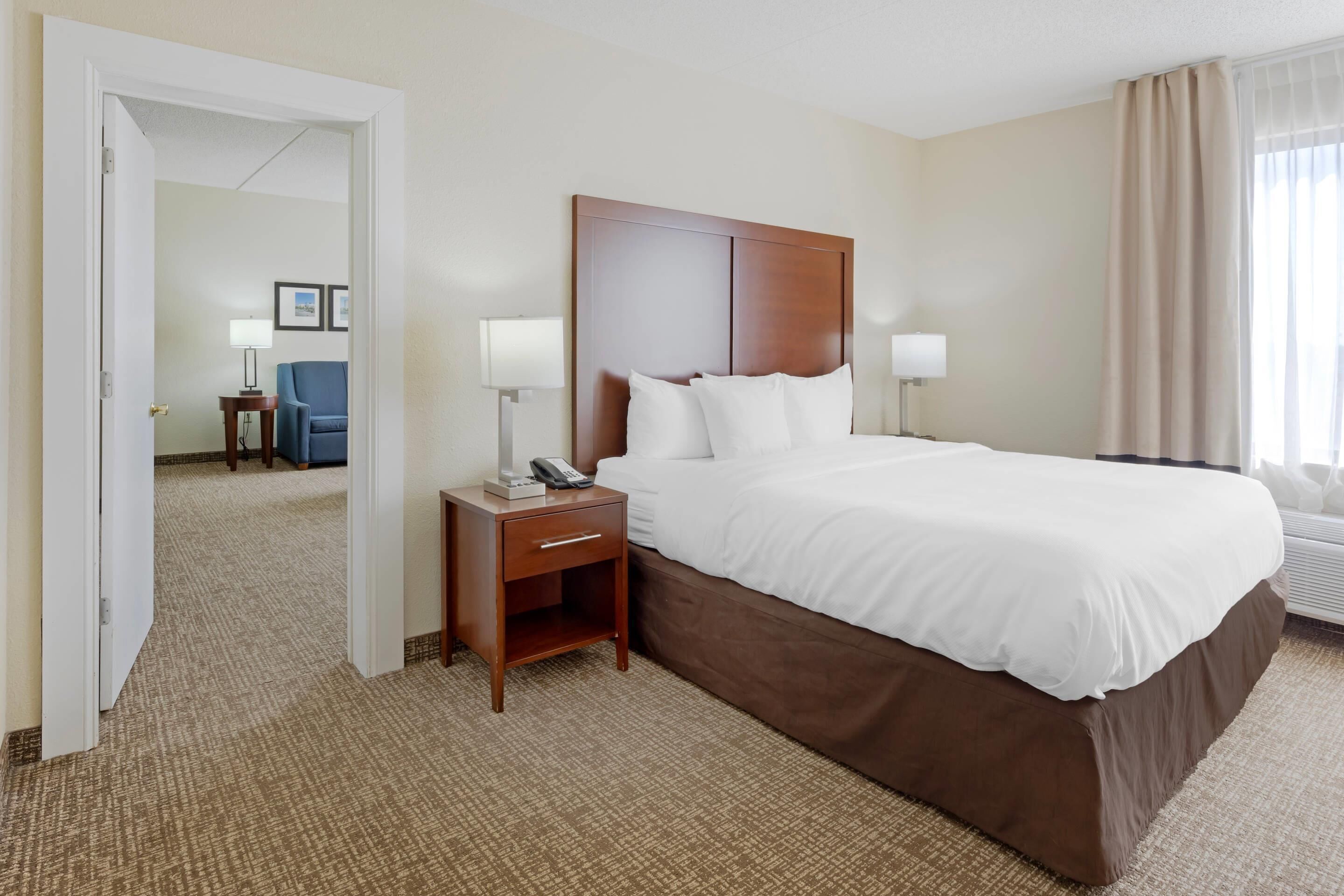 Ein Hotelzimmer mit einem großen Bett, Nachttischlampen, einem Nachttisch, einem Stuhl und einer Tür zu einem anderen Raum.