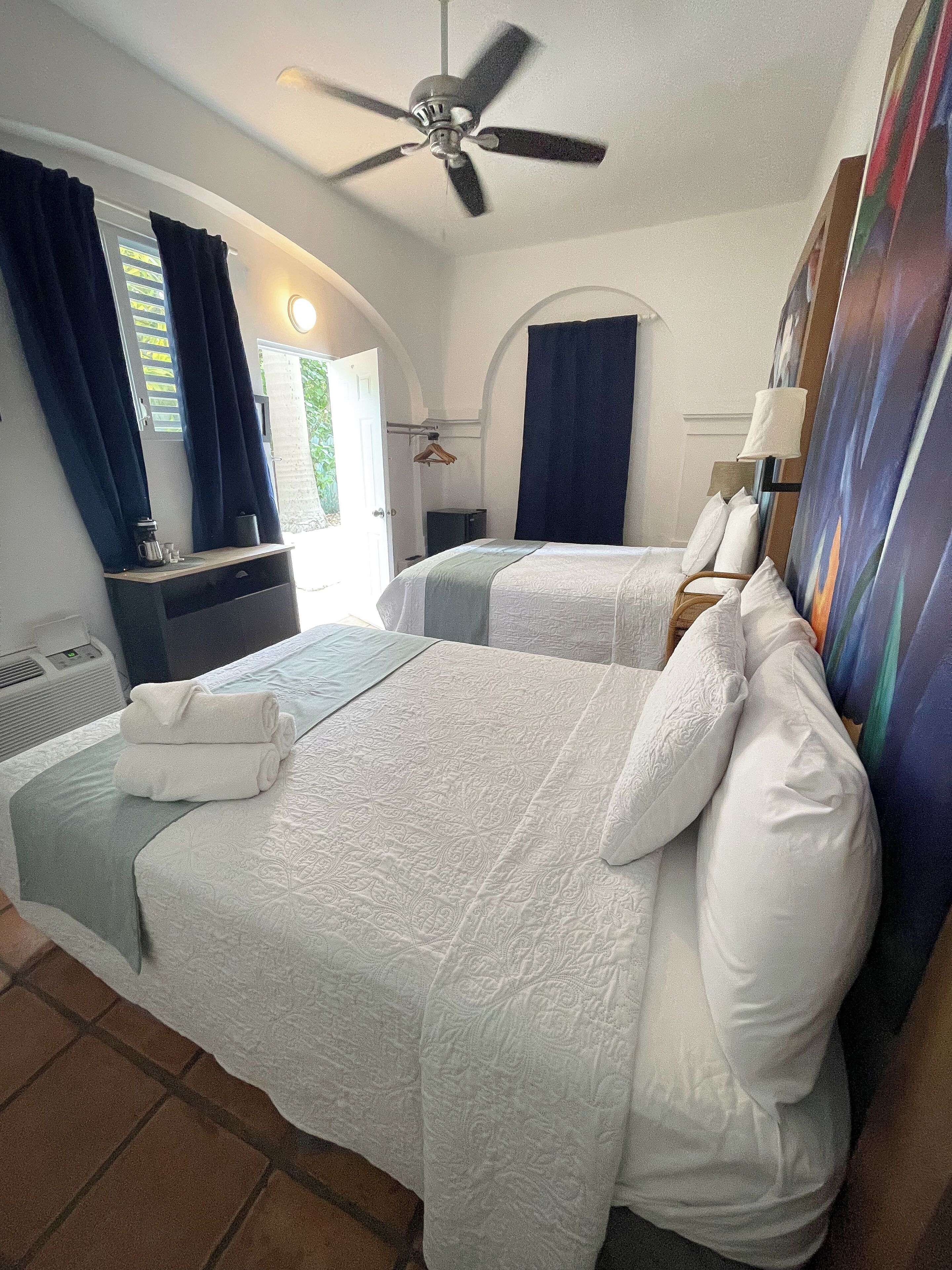Habitación de hotel con dos camas, ventilador de techo, un cuadro en la pared y ventana con cortinas.