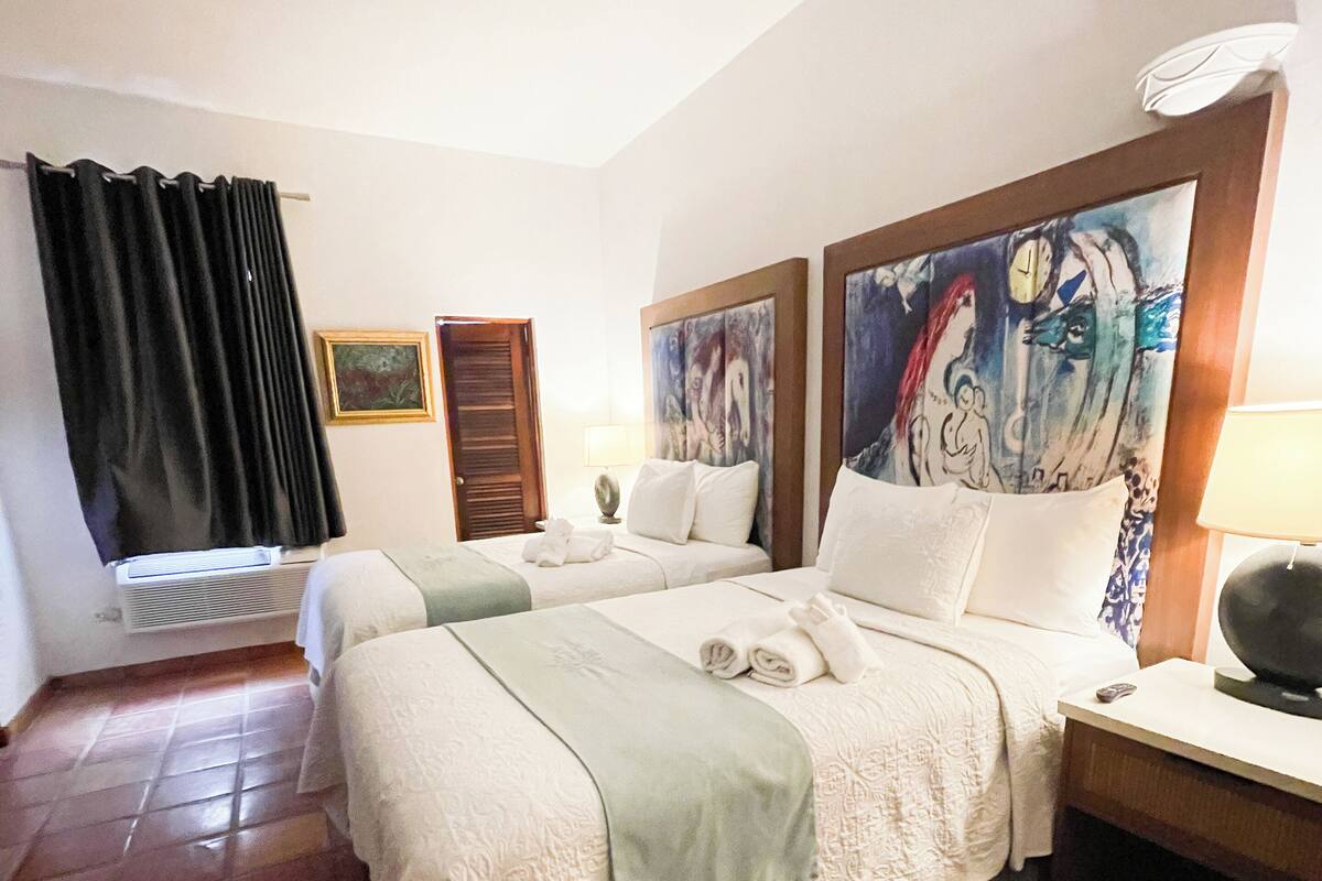A hotel room with two beds, a large painting on the wall, a desk with a lamp, and a window with curtains.