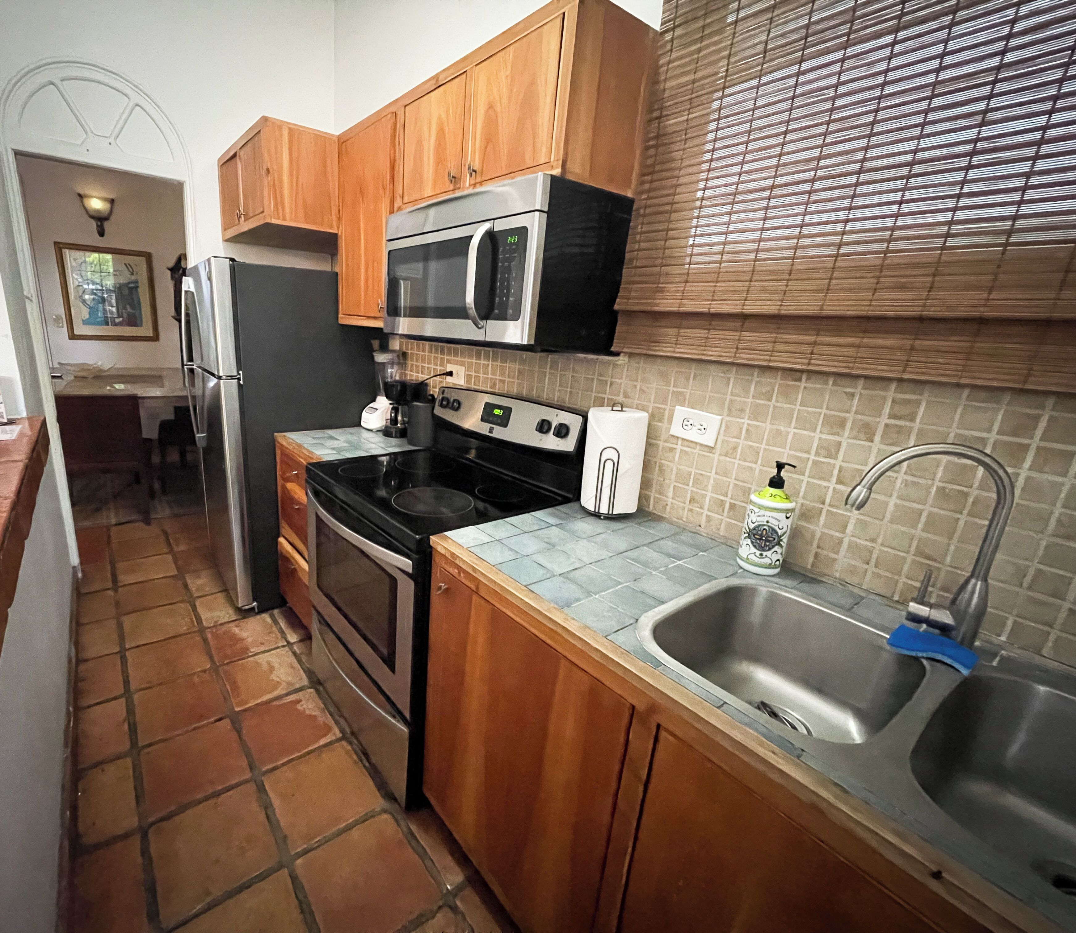 A compact kitchen with wooden cabinets, a stainless steel sink, a microwave, a stove, and a tiled backsplash.