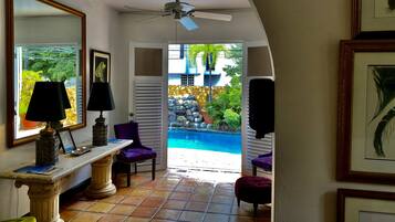 Un pasillo que conduce a una zona de piscina, con un ventilador de techo, una ventana con vistas al exterior y una mesa con una lámpara y una silla.