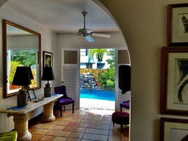 Un pasillo que conduce a una zona de piscina, con un ventilador de techo, una ventana con vistas al exterior y una mesa con una lámpara y una silla.