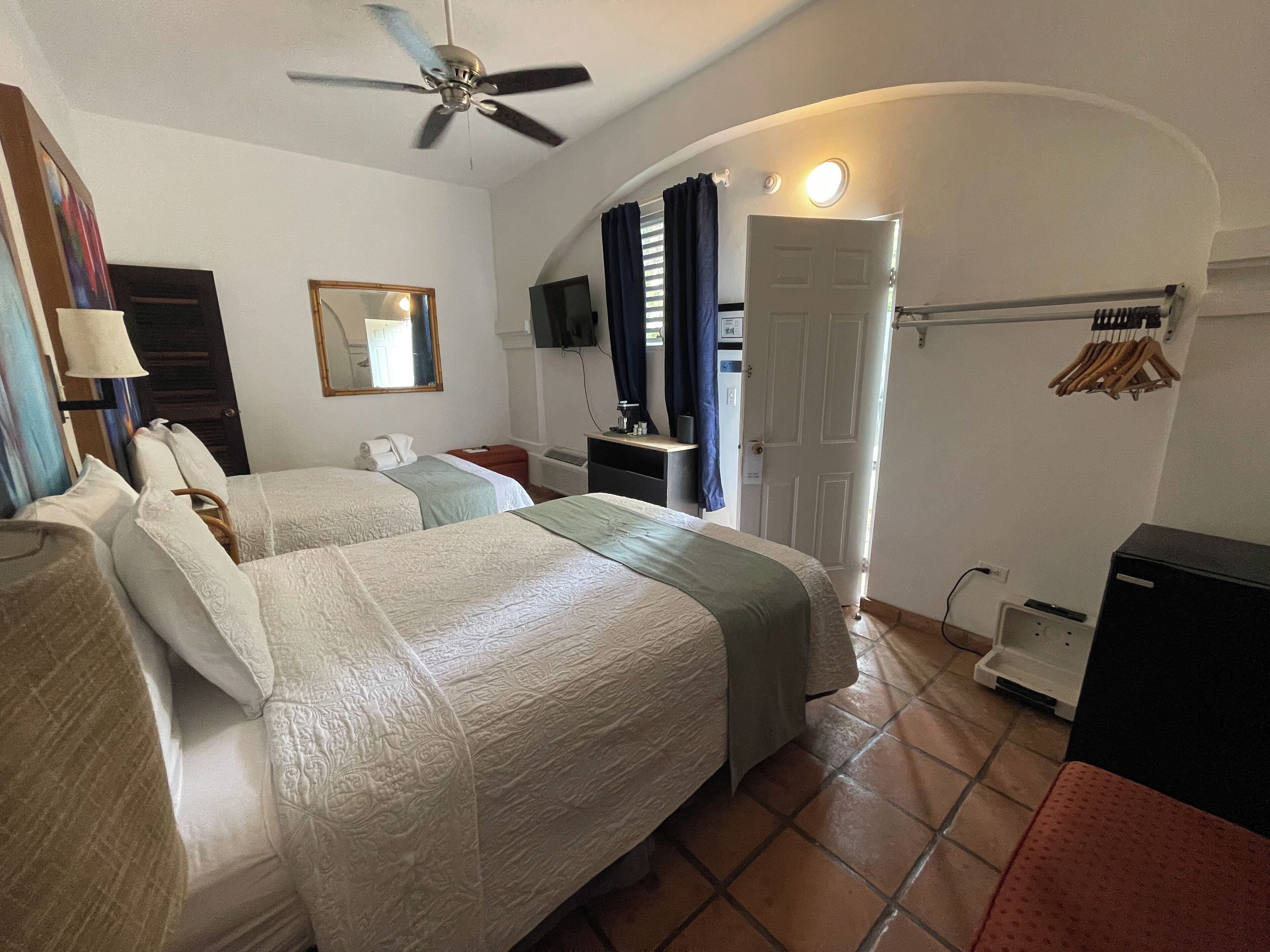 Ein Hotelzimmer mit zwei Betten, einem Deckenventilator, einem Fernseher, einem Schreibtisch und einer Badezimmertür.